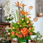 Arreglo floral exótico con aves del paraíso naranjas y rosas bicolor amarillas y naranjas, decorado con lazo naranja sobre base verde en interior de floristería