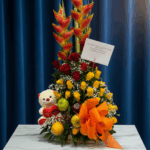 Arreglo floral vertical con rosas rojas y amarillas, heliconias naranjas, peluche blanco con corazón rojo, frutas variadas y gran lazo naranja sobre mesa de mármol