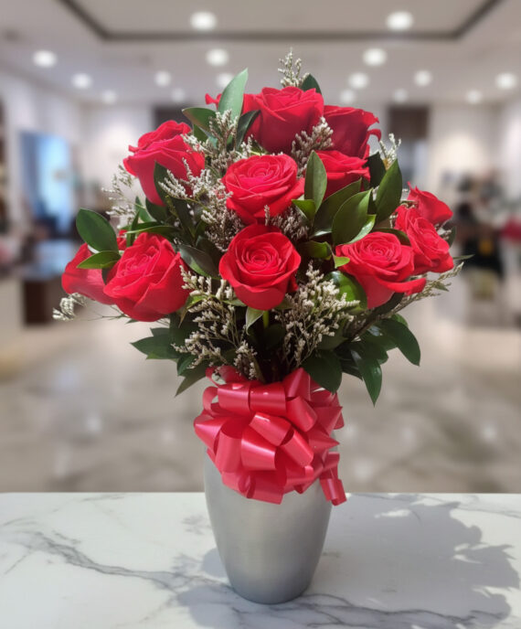 Arreglo de rosas rojas en jarrón plateado Ramo de rosas rojas con follaje verde y lazo rosa en jarrón plateado sobre mesa de mármol