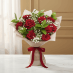 Ramo de rosas rojas frescas con follaje verde y flores blancas pequeñas, envuelto en papel beige y atado con un lazo rojo, colocado sobre una mesa de mármol blanco frente a un fondo interior cálido