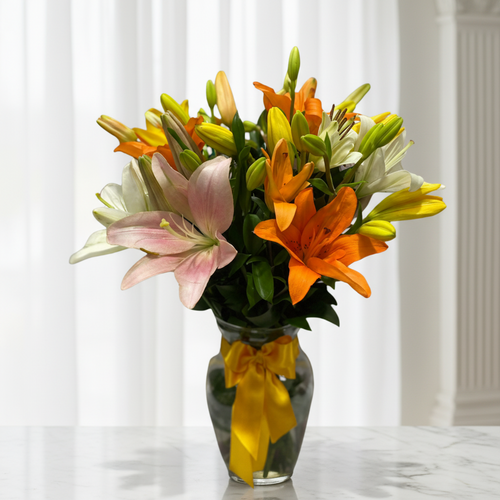Ramo de lirios de colores en jarrón de vidrio Arreglo de lirios naranjas, amarillos, blancos y rosados en un jarrón de vidrio con lazo amarillo sobre una mesa de mármol