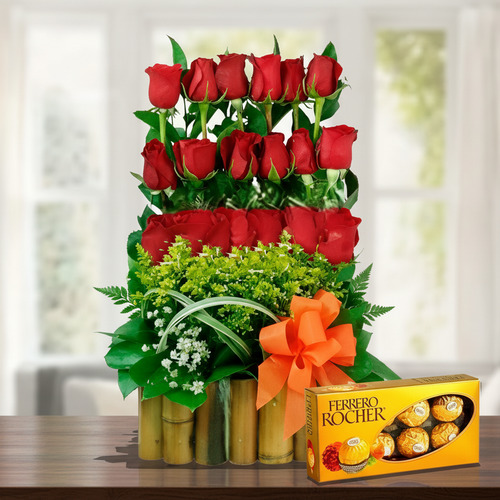 arreglo floral de rosas rojas en base de bambú con lazo naranja y caja de chocolates ferrero rocher sobre mesa