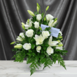 Centro de flores con rosas y lirios blancos en jarrón blanco sobre superficie de mármol, con fondo de tela gris