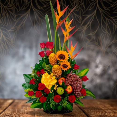 Arreglo floral tropical con frutas y lazos amarillos Arreglo floral vertical con heliconias, girasoles, rosas rojas, piñas decorativas, uvas y manzanas sobre mesa de madera con fondo oscuro