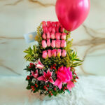 Arreglo floral vertical con rosas rosas, lirios, follaje verde, lazo rosa y globo metálico rosa sobre fondo claro