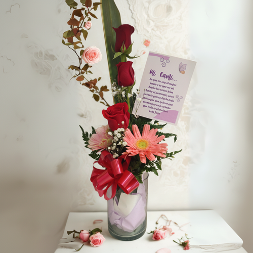 Arreglo floral en florero de vidrio transparente con rosas rojas, gerberas rosadas, follaje verde, lazo rojo y tarjeta dedicada, sobre mesa blanca con pequeñas flores dispersas