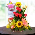 Canasta de mimbre con arreglo floral de rosas rojas, girasoles y lirios naranjas, acompañada de frutas, globo de cumpleaños en forma de estrella y tarjeta sobre una mesa redonda