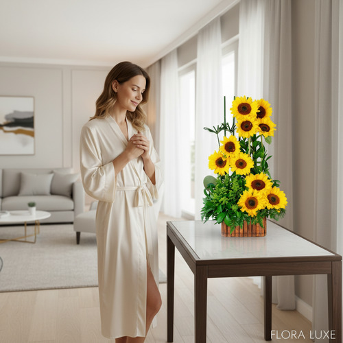 Mujer de pie con bata blanca mirando un arreglo vertical de girasoles amarillos sobre una mesa en una sala de estar luminosa y moderna