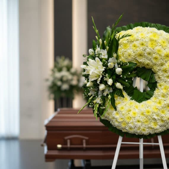Corona fúnebre Bethesda de crisantemos amarillos y flores blancas, en sala de velación moderna con ataúd desenfocado al fondo